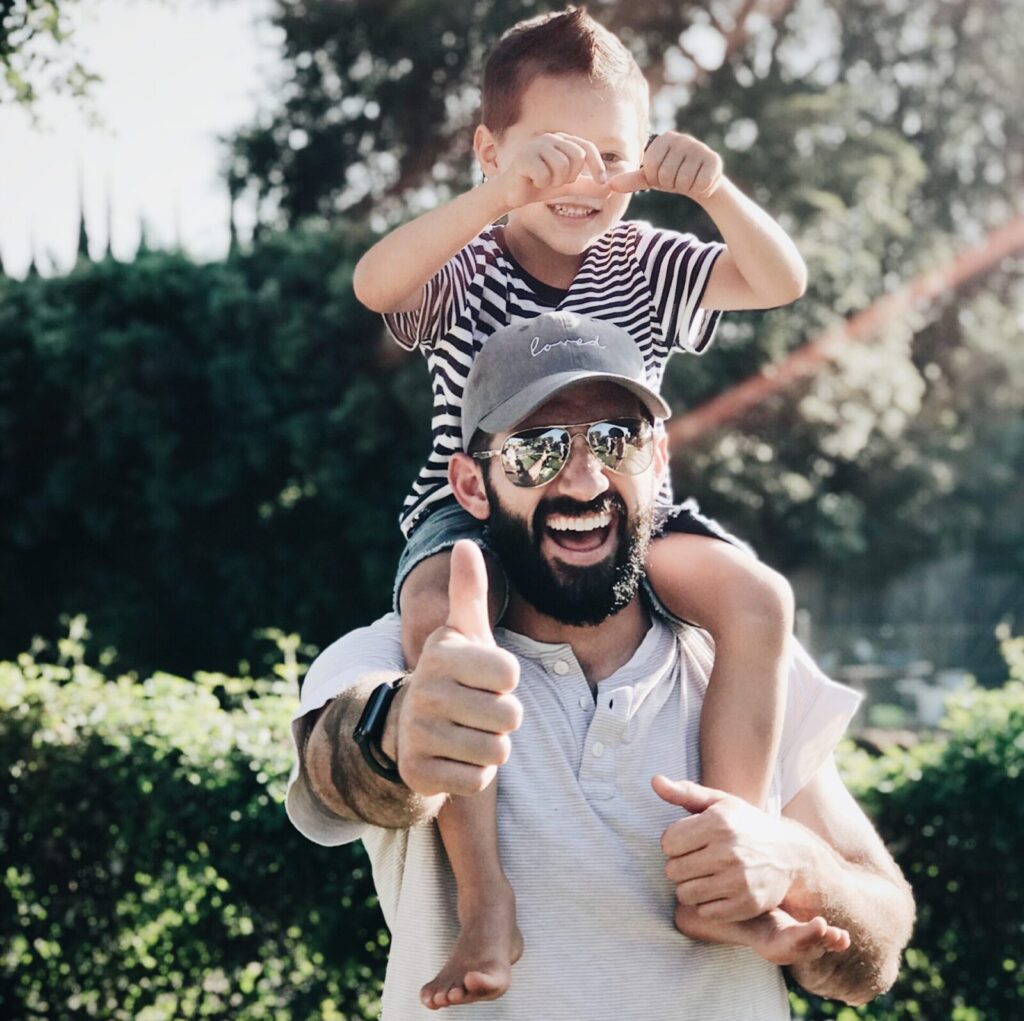 A man with a child on his shoulders. Both are very happy doing thumbs up