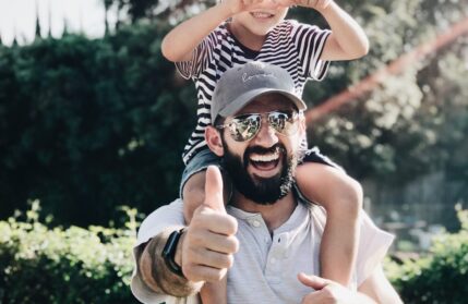 A man with a child on his shoulders. Both are very happy doing thumbs up